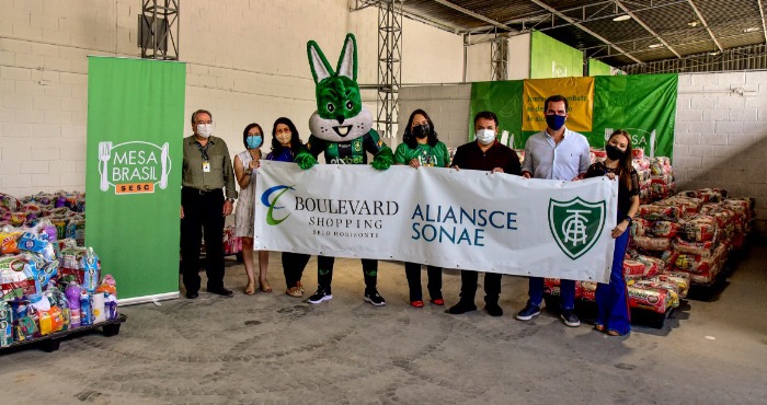 Pessoas segurando cartaz com o logo da Aliansce Sonae, Boulevard Shopping BH e Clube América. Ao fundo, doações arrecadas às vítimas das chuvas em Minas Gerais.