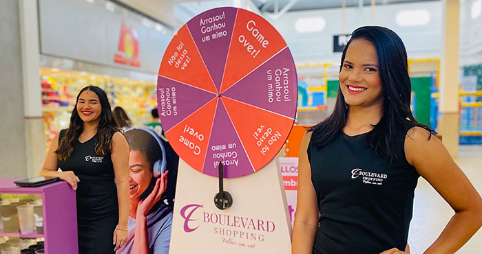 Duas promotoras ao lado da roleta de prêmios da promoção do dia do cliente ALLOS.